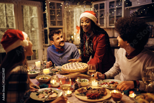 Happy black woman serving Christmas pie during dinner party with friends at dinning table.