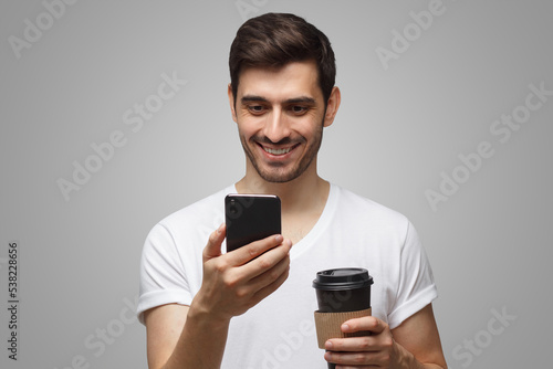 European man answering text mesage enjoying coffee from takeaway, isolated on gray photo