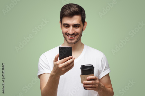 Handsome man smiling at smartphone holding takeaway coffee on green background © Damir Khabirov