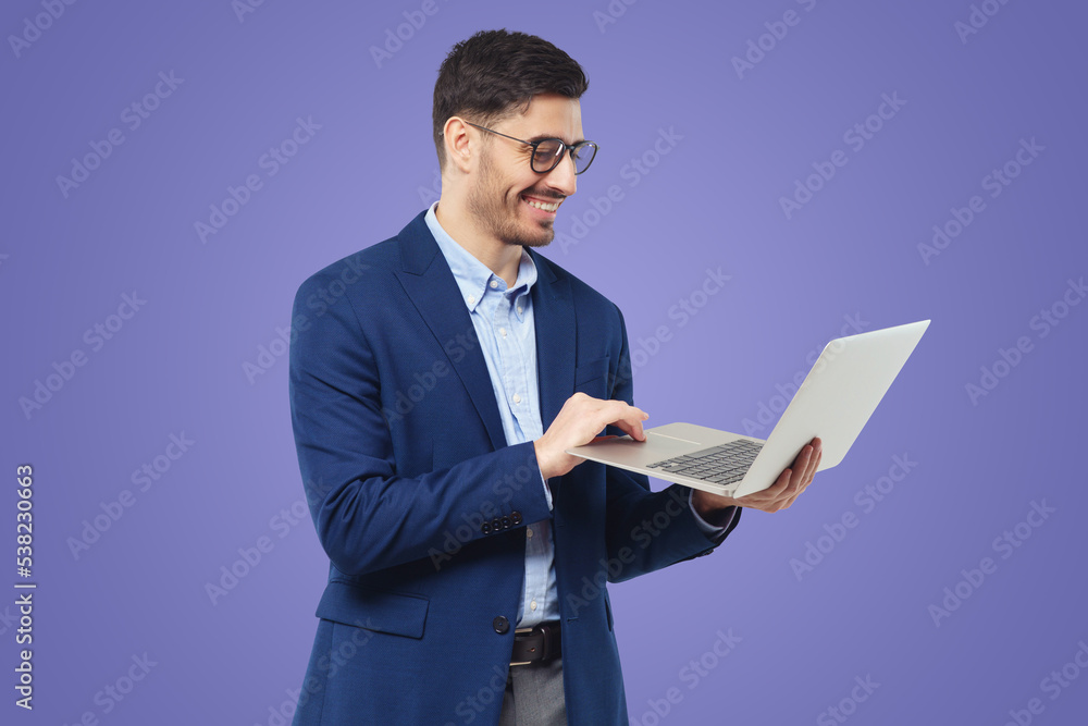 Smiling young man standing in blue suit and glasses, holding open laptop, watching funny content Smiling young man standing in blue suit and glasses, holding open laptop, watching funny content