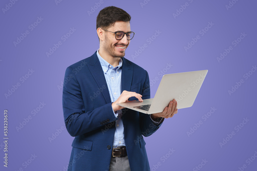 Young male teacher wearing glasses, holding open laptop, looking at screen with happy smile