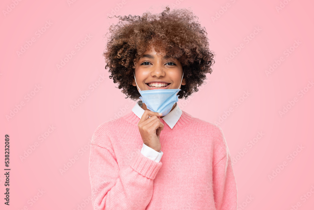 Smiling african school girl taking off face mask after the end of lockdown, isolated on pink