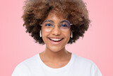 Excited laughing african teen girl in white t-shirt, eyeglasses and earphones on pink background