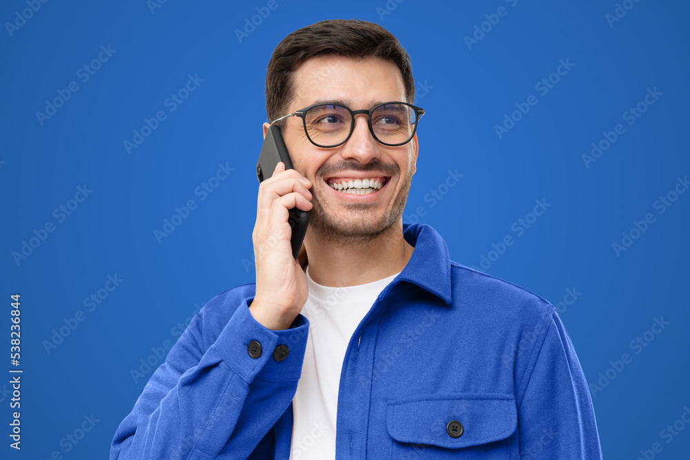 Cheerful young man in blue shirt and glasses, talking on the phone and laughing to friend's joke Cheerful young man in blue shirt and glasses, talking on the phone and laughing to friend's joke