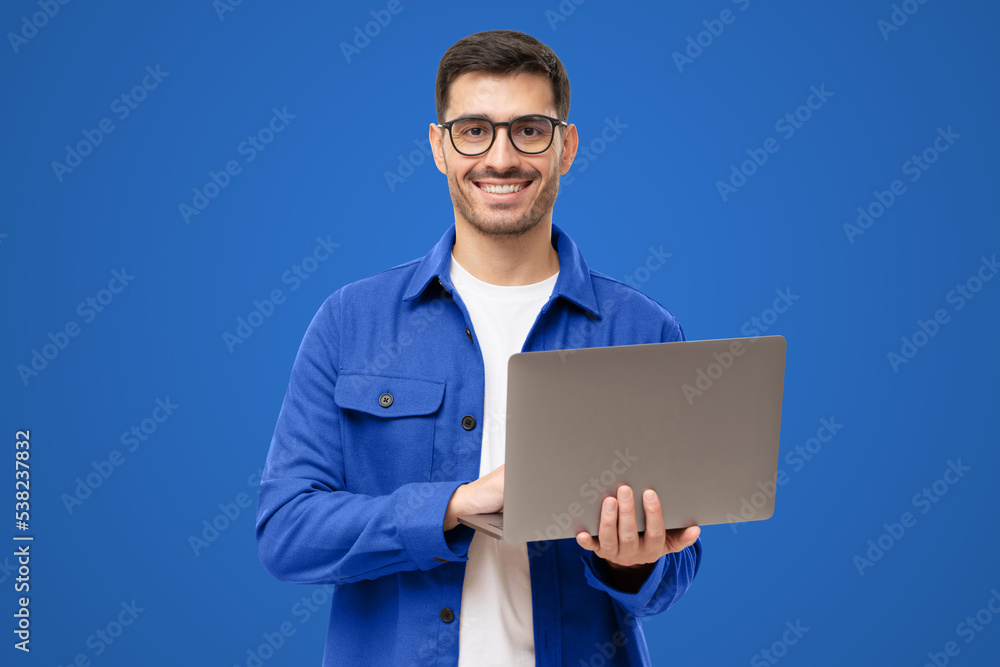 Studio portrait of young man standing holding laptop and looking at camera with happy smile Studio portrait of young man standing holding laptop and looking at camera with happy smile