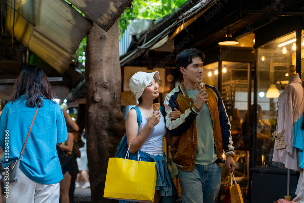 Young Asian couple enjoy and fun outdoor lifestyle shopping at street market on summer holiday vacation. Man and woman couple eating ice cream cone while walking and shopping at weekend street market.