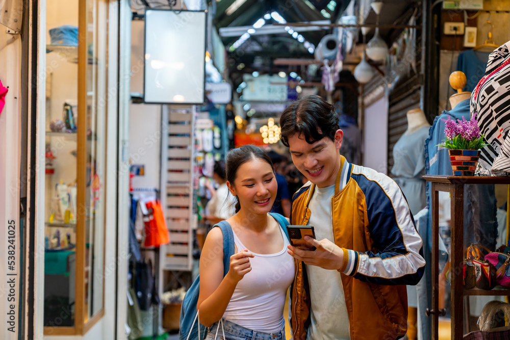 Asian couple enjoy and fun outdoor lifestyle shopping at street market on summer holiday vacation. Happy man and woman couple using mobile phone together while walking and shopping at weekend market.