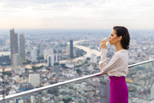  Confident Caucasian woman enjoy outdoor lifestyle drinking white wine while waiting for meeting with boyfriend at skyscraper rooftop restaurant and bar with looking to city skyline at summer sunset
