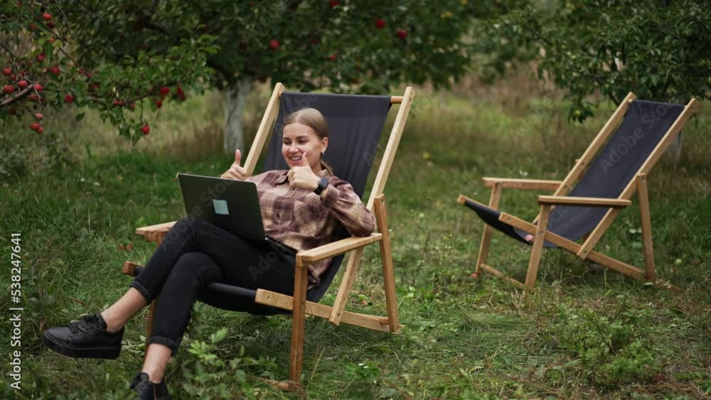 Cheerful lady having online video chat and is happy about something. Woman rejoices raising her hands and smiling happily. Nature at backdrop.