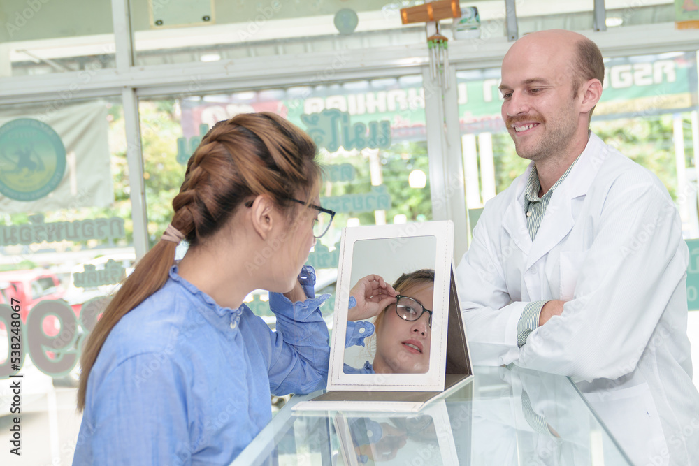 Obraz premium American man professional optician helping and selling young woman asia choosing spectacles glasses in optician store. used correct assist or defective eyesight.