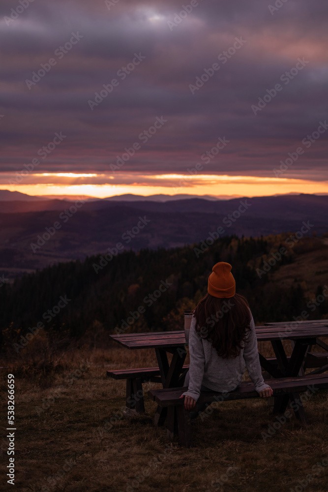 Young woman from behind is sitting at bench and look at sunset mountains view