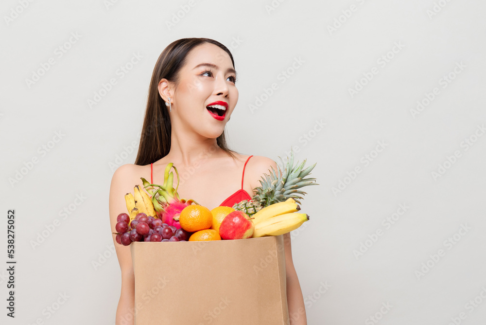 Surprised Asian woman holding paper grocery bag full of tropical fruits looking up to copy space in gray studio isolated background Surprised Asian woman holding paper grocery bag full of tropical fruits looking up to copy space in gray studio isolated background