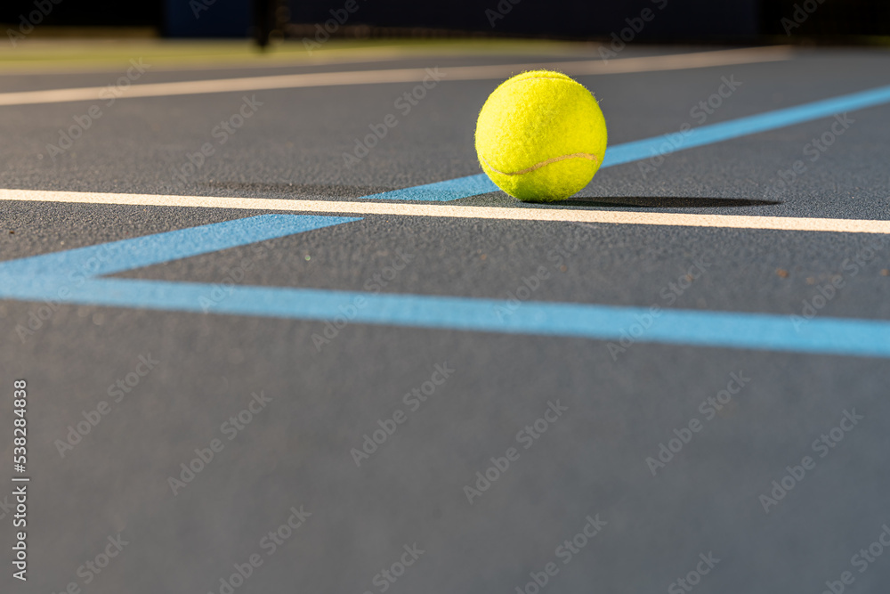 Photo taken in late evening under lights of a yellow tennis ball on blue tennis court with white line and green out of bounds.