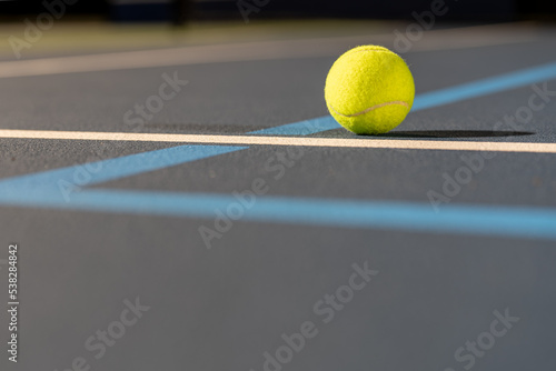 Photo taken in late evening under lights of a yellow tennis ball on blue tennis court with white line and green out of bounds. © Thomas