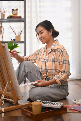 Satisfied woman sitting on floor in home studio and painting picture on canvas. Leisure activity, creative hobby and art concept