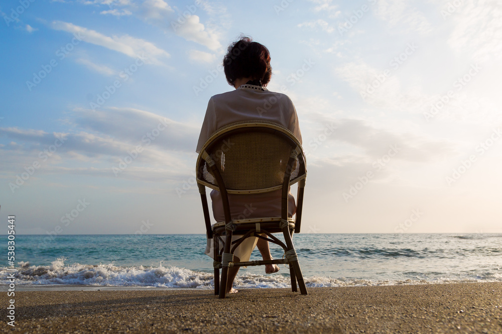 A woman in a white tunic sits on a wicker chair on the beach and looks at the sea, a view from the back.