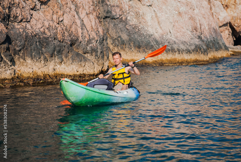A man swims in a kayak in the summer on the sea against the background of rocks