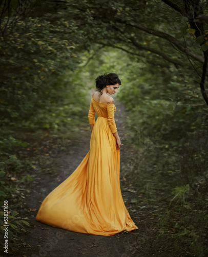 fantasy woman walking in summer nature green trees forest. Girl renaissance style princess Long vintage historical yellow dress fly in wind motion. Collected hair high hairstyle. Art photo back view 