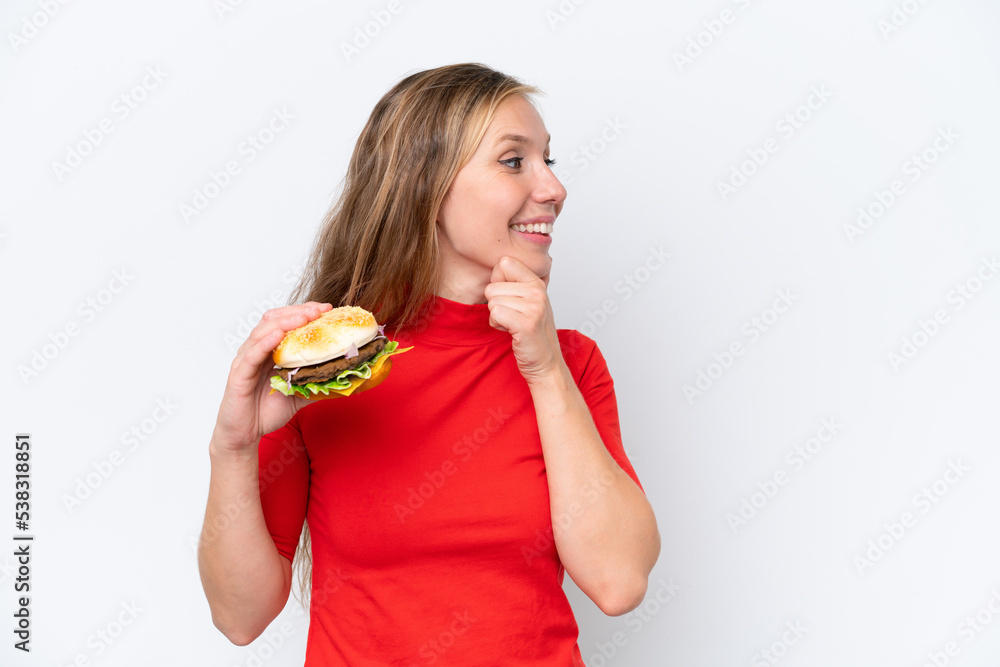 Young blonde woman holding a burger isolated on white background thinking an idea and looking side Young blonde woman holding a burger isolated on white background thinking an idea and looking side