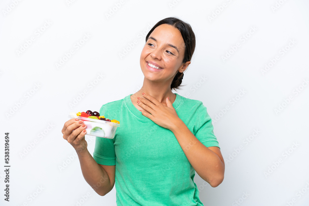 Young hispanic woman holding a bowl of fruit isolated on white background looking up while smiling Young hispanic woman holding a bowl of fruit isolated on white background looking up while smiling