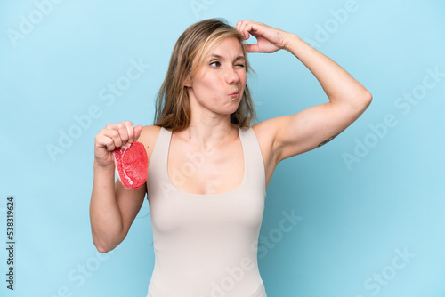 Young blonde woman holding a piece of meat isolated on blue background having doubts and with confuse face expression