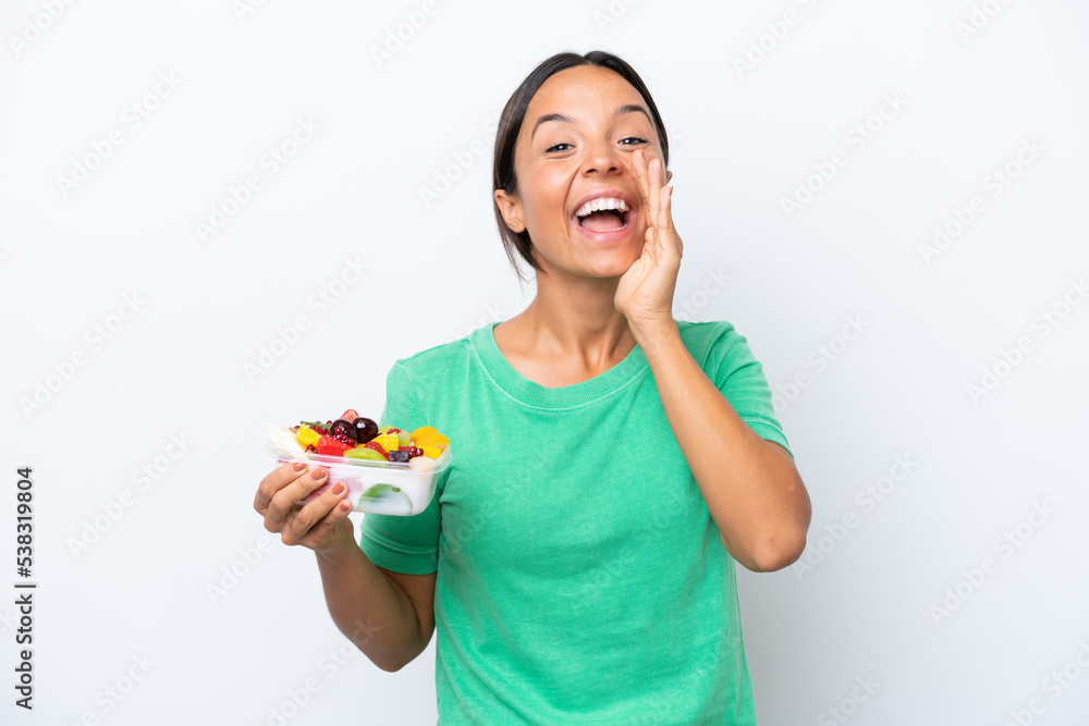 Young hispanic woman holding a bowl of fruit isolated on white background shouting with mouth wide open Young hispanic woman holding a bowl of fruit isolated on white background shouting with mouth wide open