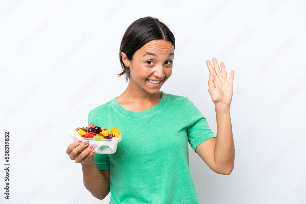Young hispanic woman holding a bowl of fruit isolated on white background saluting with hand with happy expression Young hispanic woman holding a bowl of fruit isolated on white background saluting with hand with happy expression