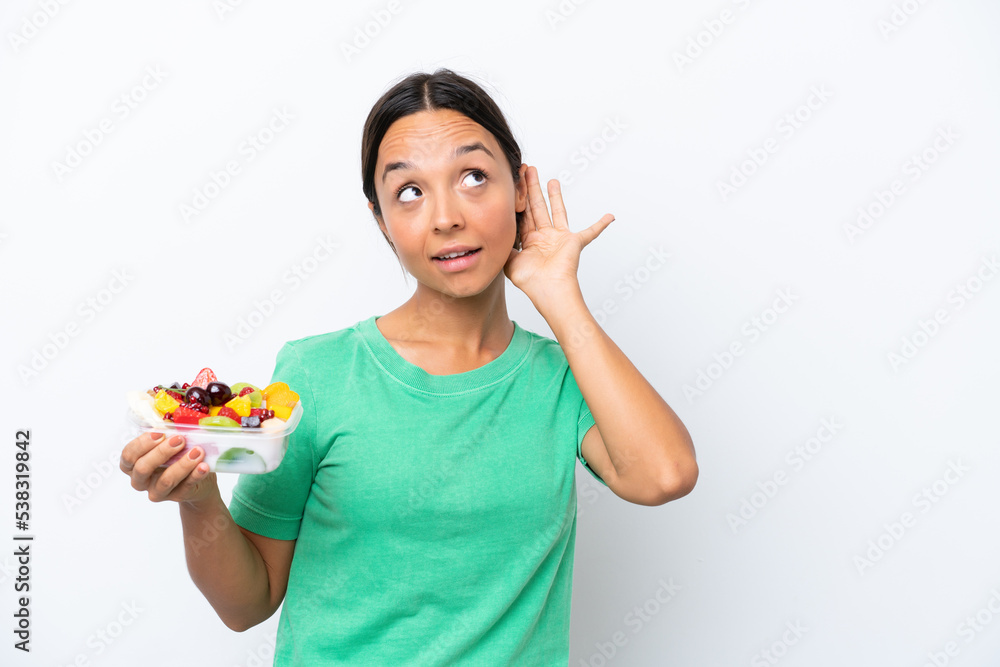 Young hispanic woman holding a bowl of fruit isolated on white background listening to something by putting hand on the ear Young hispanic woman holding a bowl of fruit isolated on white background listening to something by putting hand on the ear