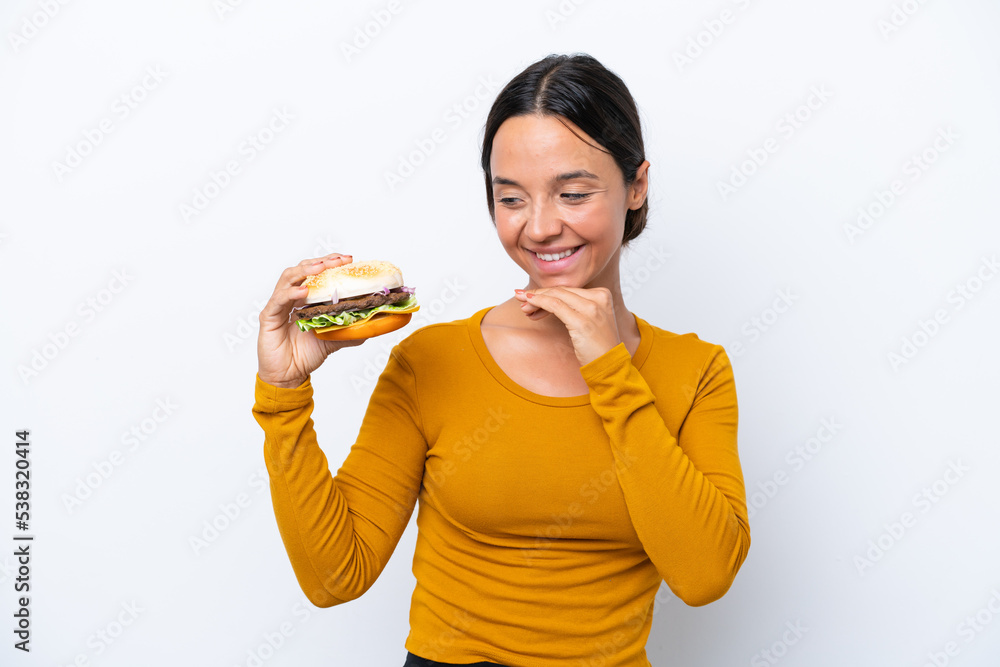Young hispanic woman holding a burger isolated on white background looking to the side and smiling Young hispanic woman holding a burger isolated on white background looking to the side and smiling