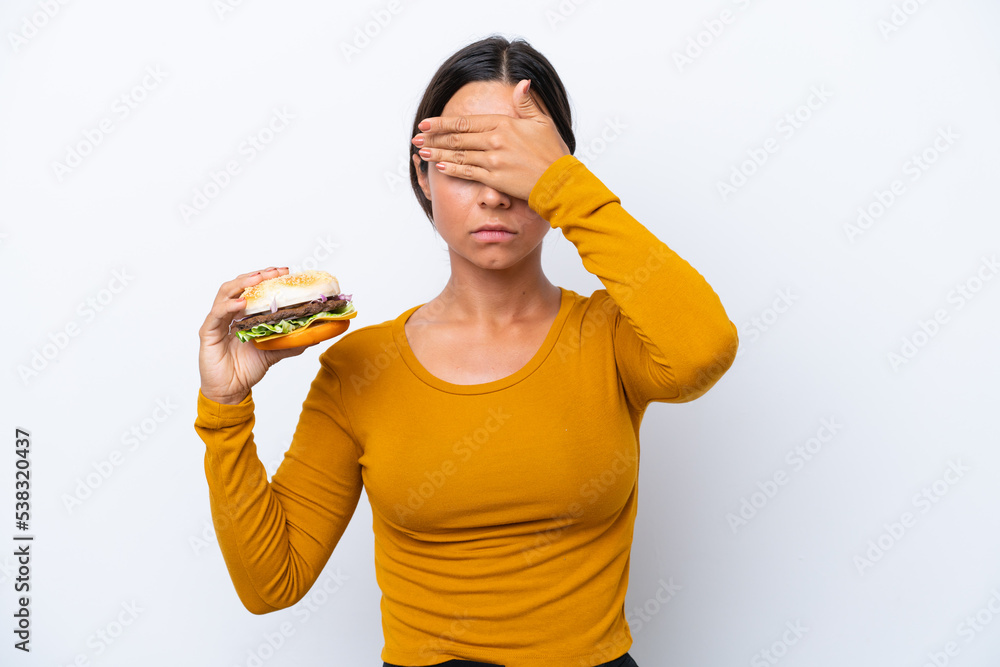 Young hispanic woman holding a burger isolated on white background covering eyes by hands. Do not want to see something Young hispanic woman holding a burger isolated on white background covering eyes by hands. Do not want to see something