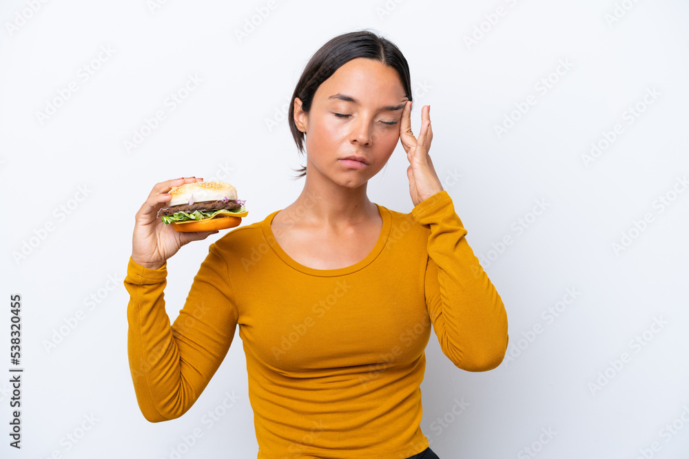 Young hispanic woman holding a burger isolated on white background with headache Young hispanic woman holding a burger isolated on white background with headache