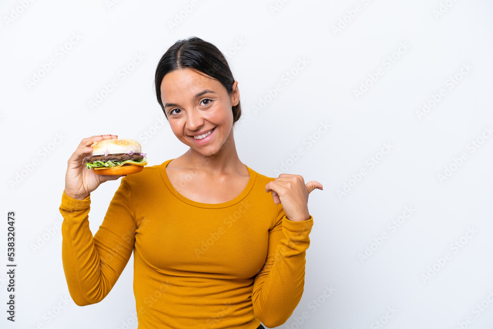 Young hispanic woman holding a burger isolated on white background pointing to the side to present a product Young hispanic woman holding a burger isolated on white background pointing to the side to present a product