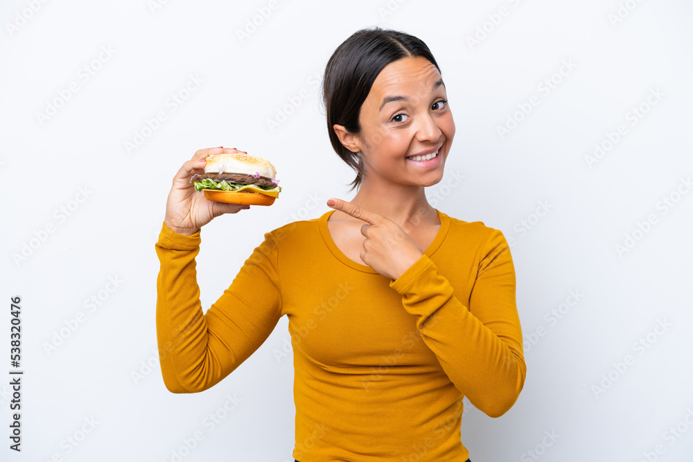 Young hispanic woman holding a burger isolated on white background pointing to the side to present a product Young hispanic woman holding a burger isolated on white background pointing to the side to present a product