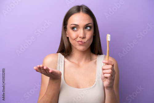 Young caucasian woman brushing teeth isolated on purple background making doubts gesture while lifting the shoulders