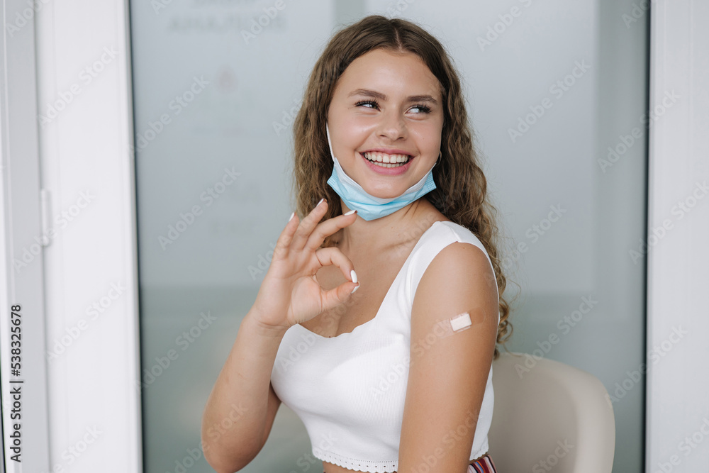 Portrait of a female smiling after getting covid-19 vaccine. Woman giving ok hand sign and showing her arm with bandage after receiving vaccination. Happy young girl with new opportunities after Portrait of a female smiling after getting covid-19 vaccine. Woman giving ok hand sign and showing her arm with bandage after receiving vaccination. Happy young girl with new opportunities after