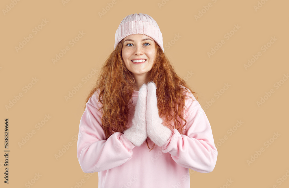Portrait of joyful woman clapping her hands joyfully in winter in anticipation of something pleasant. Cute girl with curly red hair wearing fur mittens, hat and sweatshirt isolated on beige background Portrait of joyful woman clapping her hands joyfully in winter in anticipation of something pleasant. Cute girl with curly red hair wearing fur mittens, hat and sweatshirt isolated on beige background
