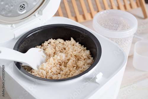 Opened electric rice cooker with cooked steaming brown rice on wooden counter-top in the kitchen photo
