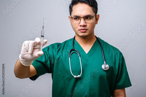 Serious asian male doctor wearing rubber gloves and scrubs, looking amazed at syringe with vaccine