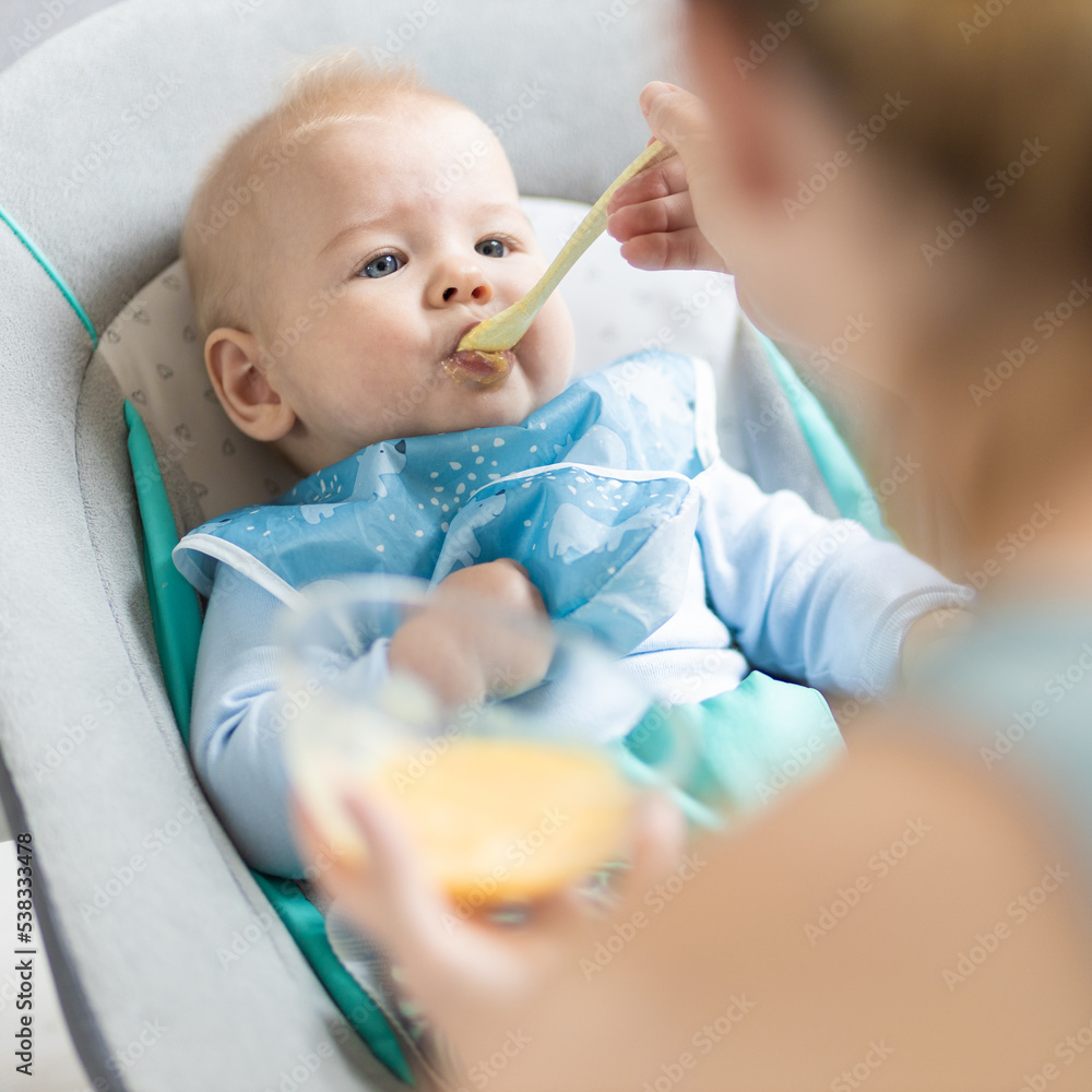 Mother spoon feeding her baby boy infant child in baby chair with fruit puree. Baby solid food introduction concept. Mother spoon feeding her baby boy infant child in baby chair with fruit puree. Baby solid food introduction concept.