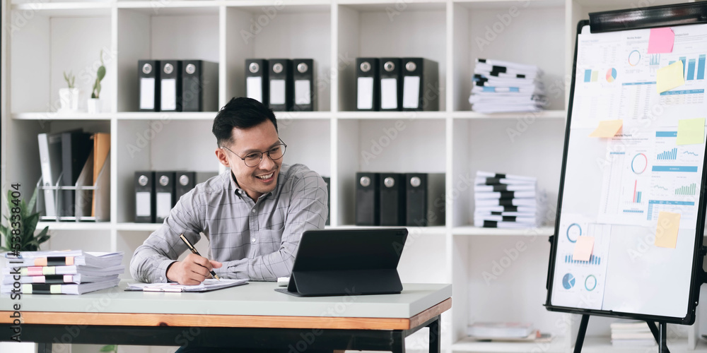 Portrait of a young Asian businessman smiling while using a laptop and writing down notes while sitting at his desk in a modern office Portrait of a young Asian businessman smiling while using a laptop and writing down notes while sitting at his desk in a modern office