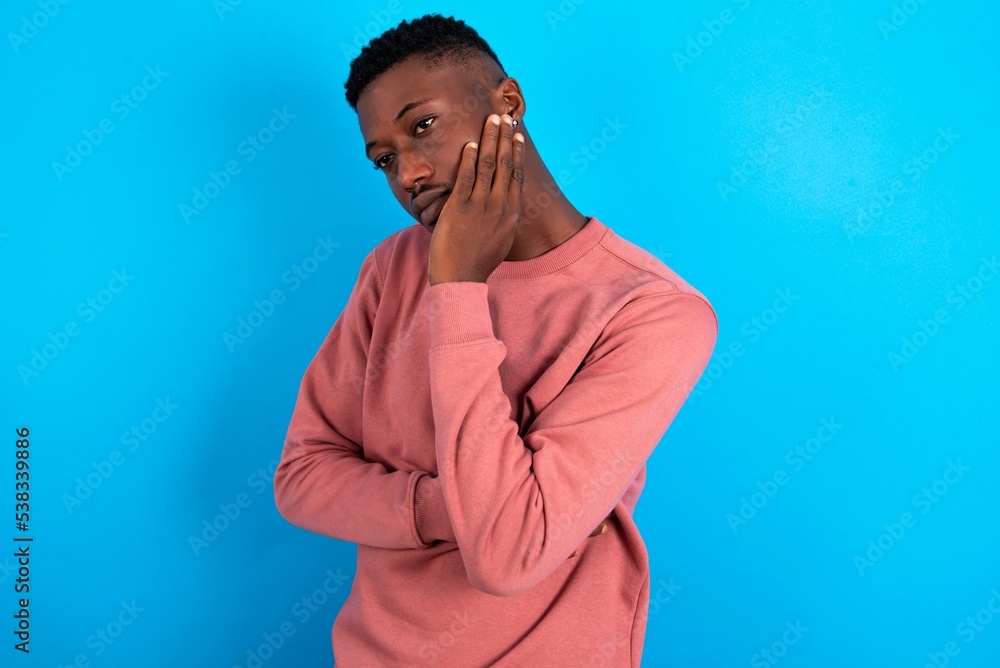 Very bored young handsome man wearing pink sweater over blue background holding hand on cheek while support it with another crossed hand, looking tired and sick,
