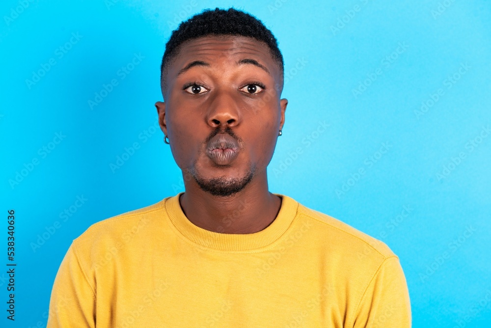 Shot of pleasant looking young handsome man wearing yellow sweater over blue background, pouts lips, looks at camera, Human facial expressions Shot of pleasant looking young handsome man wearing yellow sweater over blue background, pouts lips, looks at camera, Human facial expressions