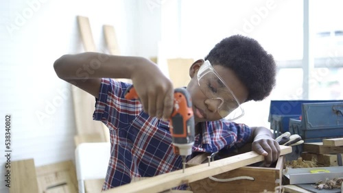 A young african boy wearing shirt and using screwdriver and try to screw the wood piece in the classroom. Smiling kid carpenter happy working with wood and sandpaper, preschool and learning concept.
 photo