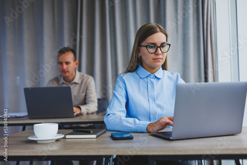 Focused woman in casual clothes typing on a netbook while working on a new project while sitting at a desk in a modern workspace. Business lady in a jacket and glasses. Remote work