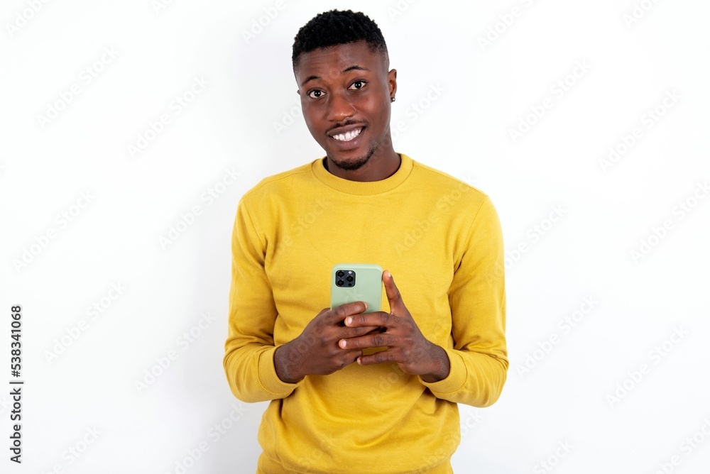 Smiling young handsome man wearing yellow sweater over white background friendly and happily holding mobile phone taking selfie in mirror. Smiling young handsome man wearing yellow sweater over white background friendly and happily holding mobile phone taking selfie in mirror.