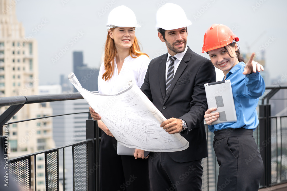 Engineer man and architect coworker standing outdoors on rooftop of construction site looking at blueprint for apartment, housing development. Maintenance building contractor discussion with teamwork Engineer man and architect coworker standing outdoors on rooftop of construction site looking at blueprint for apartment, housing development. Maintenance building contractor discussion with teamwork
