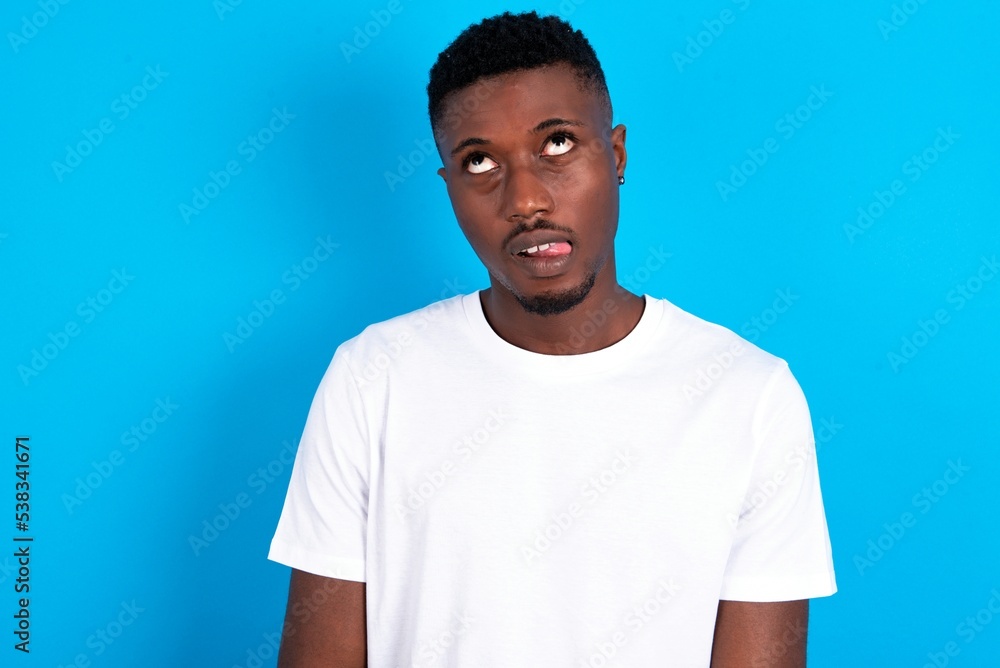Funny young handsome man wearing white T-shirt over blue background makes grimace and crosses eyes plays fool has fun alone sticks out tongue. Funny young handsome man wearing white T-shirt over blue background makes grimace and crosses eyes plays fool has fun alone sticks out tongue.