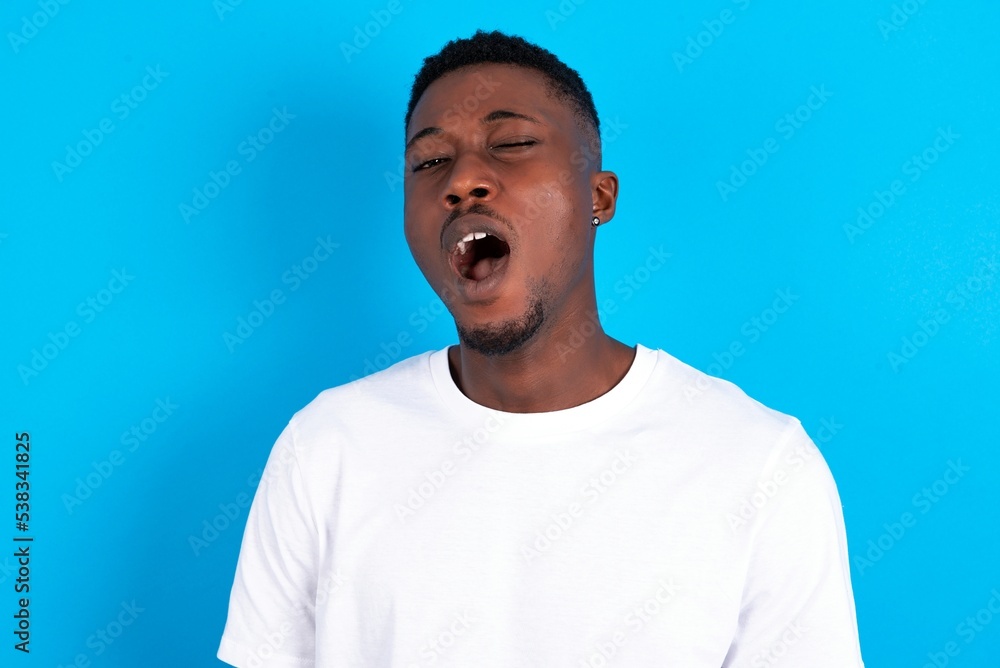 young handsome man wearing white T-shirt over blue background yawns with opened mouth stands. Daily morning routine
