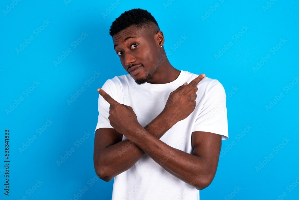 young handsome man wearing white T-shirt over blue background crosses arms and points at different sides hesitates between two items or variants. Needs help with decision young handsome man wearing white T-shirt over blue background crosses arms and points at different sides hesitates between two items or variants. Needs help with decision