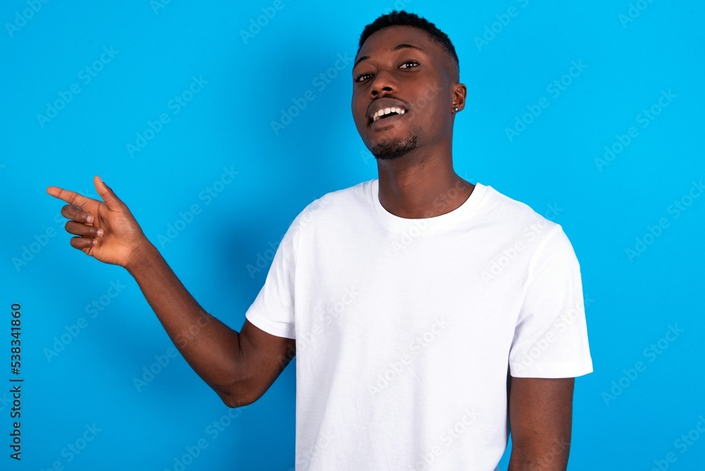 young handsome man wearing white T-shirt over blue background laughs happily points away on blank space demonstrates shopping discount offer, excited by good news or unexpected sale. young handsome man wearing white T-shirt over blue background laughs happily points away on blank space demonstrates shopping discount offer, excited by good news or unexpected sale.